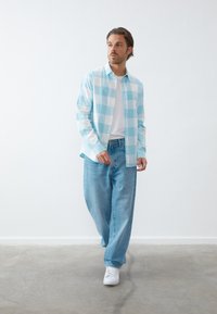Hellblau-weiß kariertes Hemd, knöpfbar, über einem weißen T-Shirt getragen. Kombiniert mit locker sitzenden hellblauen Jeans und weißen Turnschuhen.