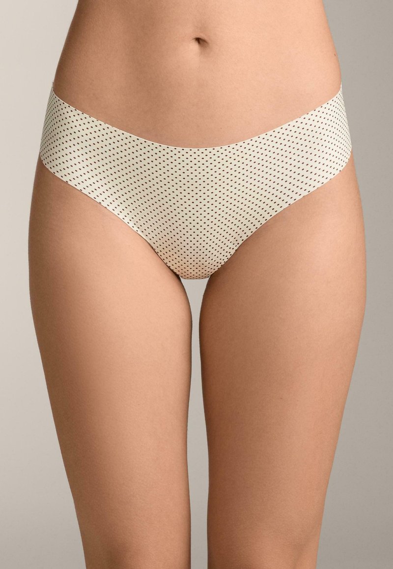 Femme portant des sous-vêtements beiges taille haute à petits pois noirs, debout devant un fond neutre.
