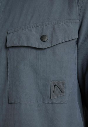 Tmavosivá látková náprsní kapsa s chlopňou upevnenou čiernym cvokovým gombíkom a malou textúrovanou logovou nášivkou v dolnom rohu.