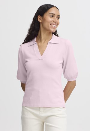 Femme aux longs cheveux foncés sourit, portant un haut rose clair à manches courtes avec un col en V et un pantalon blanc sur un fond uni.
