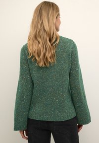 Teal ulltröja med en texturerad yta, vida ärmar och en avslappnad passform. Har en rund halsringning och subtila inslag av orange och gult.