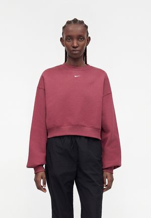 Mujer con el pelo trenzado, vistiendo una sudadera holgada de Nike color granate y pantalones negros, de pie con los brazos relajados contra un fondo blanco liso.