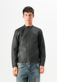 Jeune homme portant une veste en cuir noire zippée avec des épaules matelassées et un jean bleu, se tenant devant un fond blanc uni.