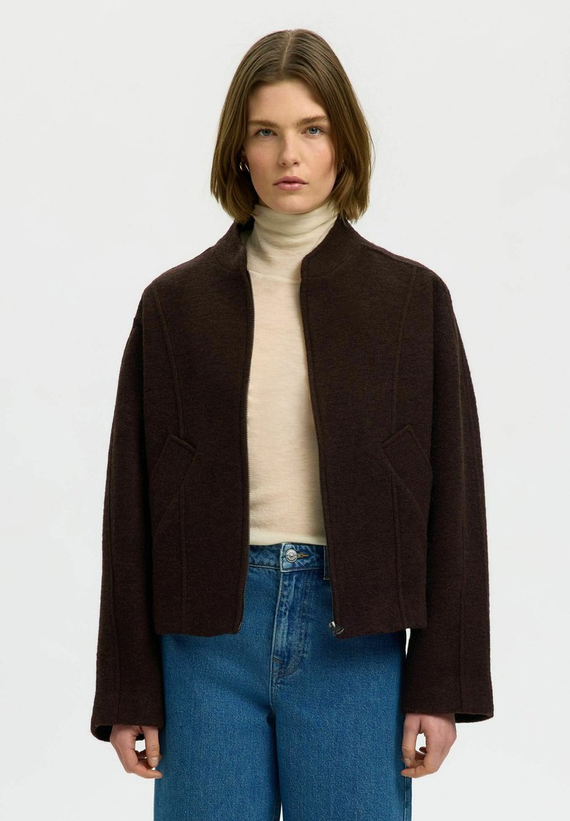 Femme portant une veste zippée marron foncé sur un col roulé beige et un jean bleu, debout devant un fond clair uni.