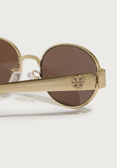 Kružne sunčane naočale s smešnim lećama i zlatnim metalnim okvirom. Ručke sadrže kružni detalj s logotipom Tory Burch. Glatka tekstura.