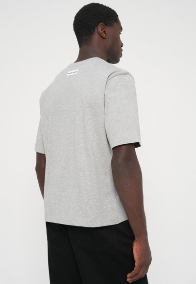 Sivá tričko s krátkym rukávom z mäkkého materiálu, má rovný spodný lem a malú nášivku pri výstrihu. Nosí sa s čiernymi nohavicami.