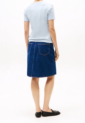 Jeansowa spódnica w kroju A, o średniej długości, w ciemnoniebieskim kolorze, z dwiema tylnymi kieszeniami i widoczny odszyciem. Połączona z jasnoniebieską koszulką.