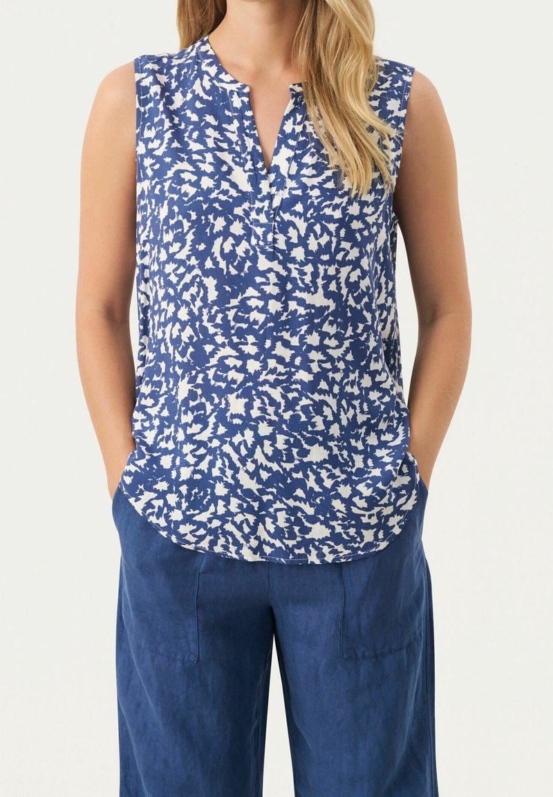 Femme portant un chemisier sans manches bleu et blanc à motifs et un pantalon bleu ample, les mains dans les poches, sur fond uni.