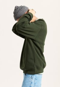 Sudadera verde oscuro de manga larga, con puños acanalados y una etiqueta en la costura inferior. Combinada con jeans azul claro y un gorro gris.