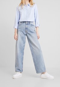 Hellblaues Button-up Hemd, lockere Passform, getragen mit hellen, weit geschnittenen Jeans und weißen Sneakers. Glattes Material mit aufgerollten Ärmeln.