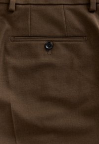 Pantalon en tissu marron avec une poche passepoilée unique à l'arrière, fermée par un bouton noir, et des passants de ceinture visibles à la taille.