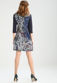 Robe à motifs avec manches trois-quarts ; présente un design bleu et floral, un tissu texturé, et une jupe évasée, complétée par des talons hauts.
