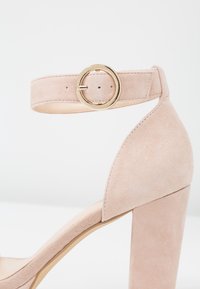 Svetle růžové semišové sandály na vysokém podpatku s širokým podpěrou a řemínkem kolem kotníku, který má kulatou zlatou sponu.