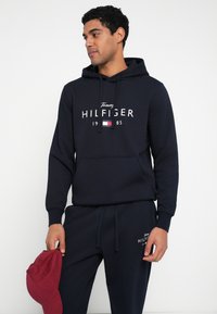 Granatowa bluza z kapturem wykonana z bawełnianej mieszanki, z przednią kieszenią, ozdobiona białym logo i nadrukiem z rokiem. W komplecie spodnie do kompletu i czerwona czapka.