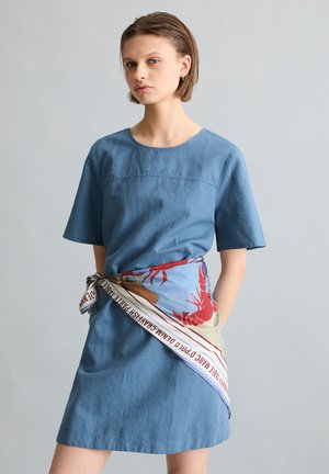 Młoda kobieta w niebieskiej sukience z krótkim rękawem z kolorowym szalikiem w nadruk homara przewiązanym wokół talii na jednolitym tle.