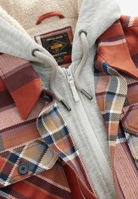Geruite jas met grijze fleece voering. Heeft rode, blauwe en beige ruiten, ritssluiting, capuchon met trekkoord en metalen accenten. Zachte textuur.