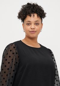 Zwarte top met een gladde stoffen body en doorschijnende puffmouwen met fluwelen stippen, ronde halslijn en een relaxed fit.