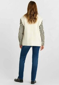 Gilet di maglia crema oversize indossato sopra una maglietta a maniche lunghe a righe bianche e nere, abbinato a jeans blu e scarpe nere, visto da dietro.