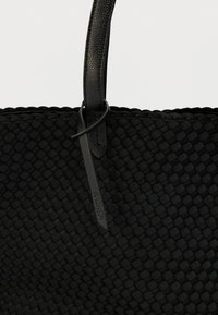 Μαύρη υφαντή τσάντα ώμου με υφή στην επιφάνεια. Λεπτές λαβές από λείο δέρμα και ετικέτα που αναγράφει το εμπορικό σήμα. Απλός, λειτουργικός σχεδιασμός.