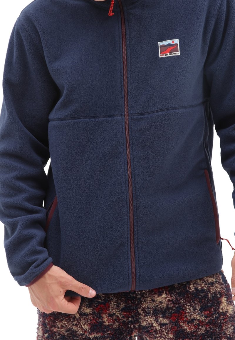 Personne portant une veste polaire bleu marine zippée avec un petit logo rectangulaire rouge et blanc sur la poitrine et un pantalon à motifs.