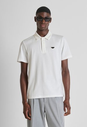 Uomo con occhiali da sole rettangolari neri, polo bianca a maniche corte con piccolo logo sul petto e pantaloni grigio chiaro a pieghe, in piedi.