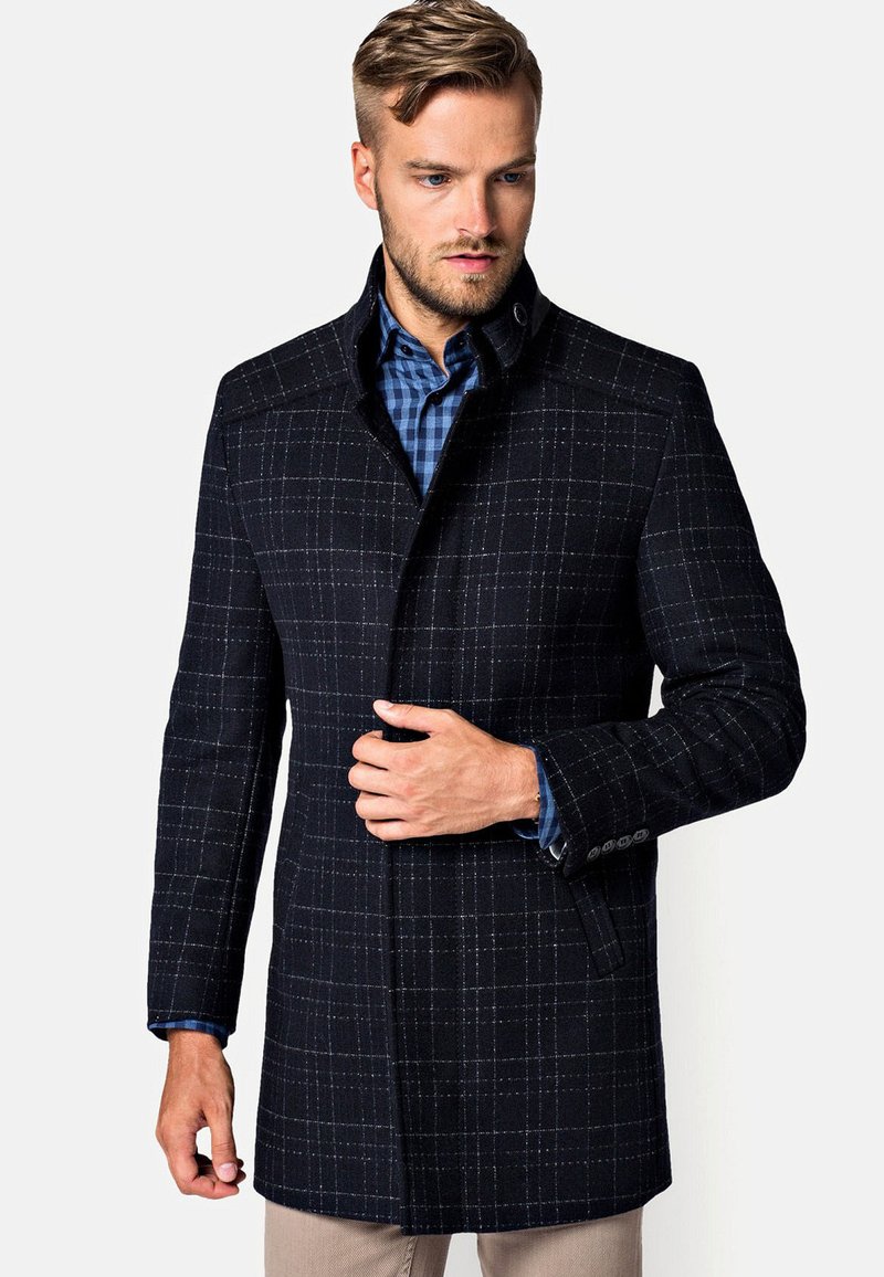 Granatowy płaszcz w kratę z wełny z wyraźnym kołnierzem, guzikowymi akcentami na rękawach i dopasowanym krojem. Pod spodem kontrastowa niebieska koszula w kratę.
