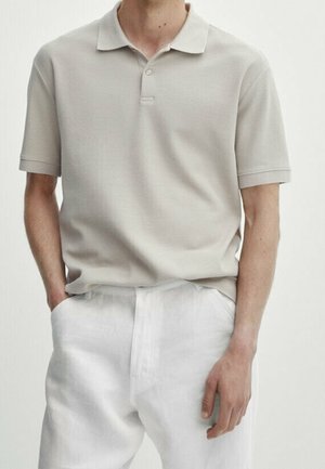 Mężczyzna w jasnoszarej koszulce polo z krótkim rękawem, włożonej do białych spodni z wysokim stanem, trzyma jedną rękę w kieszeni.