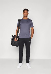 Szürke sportos póló, fekete logós melegítőnadrág, fekete hátizsákot tartva. Fehér sportcipő. Egyszerű, testhez álló, sportos megjelenés.