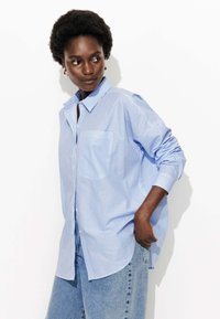 Chemise à rayures bleues et blanches avec un col, dotée d'une poche et d'une coupe ample, assortie à un jean bleu taille haute.