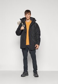 Giacca nera isolata con cappuccio, indossata sopra una maglietta arancione. Pantaloni scuri e scarpe nere completano l'outfit. Il modello tiene in mano una borsa.