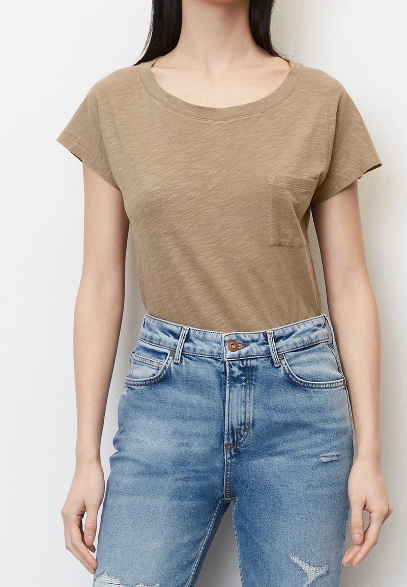 Femme portant un t-shirt marron clair à manches courtes avec une poche poitrine, associé à un jean délavé bleu clair taille haute.