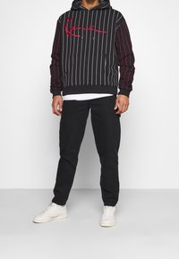 Musta huppari, jossa valkoiset pystysuorat raidat ja punainen kirjailu, edessä kengurutasku ja huppu, yhdistetty mustiin housuihin ja valkoisiin lenkkareihin.