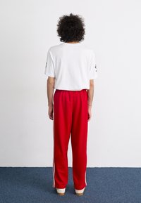 Hvit bomull t-skjorte med korte ermer, kombinert med vide røde joggebukser med hvite sidestriper, og beige sko. Bakside.