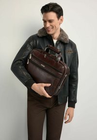 Brązowa skórzana aktówka z wieloma zamykanymi na zamek przegródkami, solidnymi uchwytami i gładką teksturą; trzymana przez osobę w ciemnej kurtce.