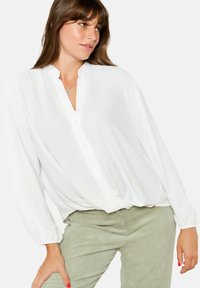 Blusa bianca com vestibilità ampia, colletto alla mandarino, chiusura frontale con bottoni e lunghe maniche voluminosi. Abbinata a pantaloni verdi chiari con texture.