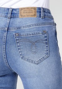 Blue denim jeans featuring a brown label with "KOROSHI" text, accent stitching on the back pocket, and a fitted design.
