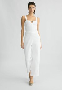 Combinaison blanche avec décolleté carré, détail de taille tressée, pantalon large, et texture légèrement brillante ; assortie avec des talons hauts à motifs.