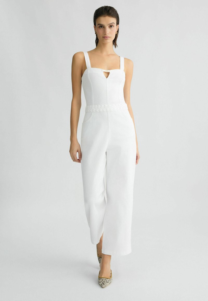 Combinaison blanche avec décolleté carré, détail de taille tressée, pantalon large, et texture légèrement brillante ; assortie avec des talons hauts à motifs.