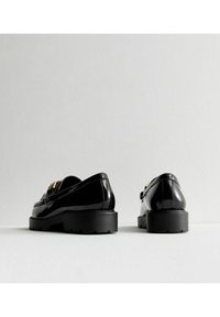 Mocasines de charol negro con suela gruesa negra, que presentan un detalle de hebilla dorada y costuras texturizadas en la parte superior.