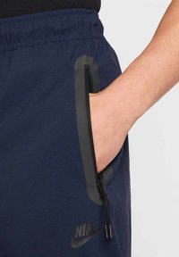Marine sportshorts met een zwart Nike-logo, een ritszak aan de zijkant met grijze accenten, een elastische tailleband en een gladde stofstructuur.