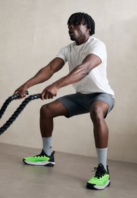 Atleta esegue uno squat utilizzando corde da combattimento nere, indossando una maglietta bianca aderente, pantaloni corti grigi e sneakers nere/verde con dettagli testurizzati.