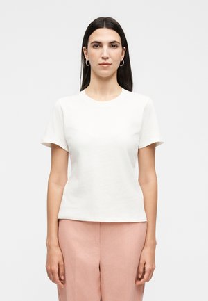 Kvinde med langt mørkt hår iført hvid kortærmet skjorte, lyserøde bukser og ringformede øreringe, stående mod en ensfarvet hvid baggrund.