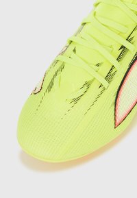 Felgele voetbalschoen met een gestructureerd bovenwerk, voorzien van zwarte patronen en roze accenten, vastgemaakt met veters in een gestroomlijnd ontwerp.