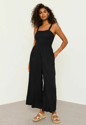 Göndör hajú nő, ujjatlan fekete overált visel, amelynek bő szárú nadrágja van, zöld fülbevalókat, karkötőket és virágmintás csattos szandált.