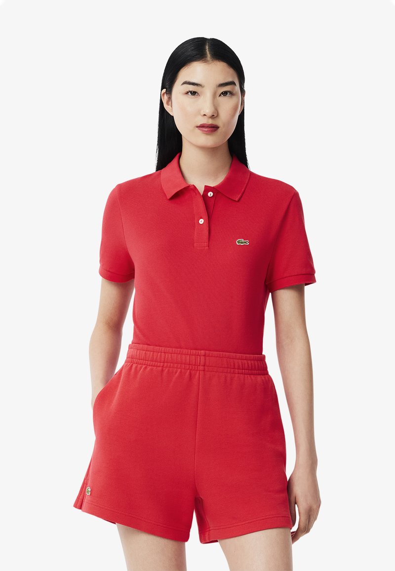 Punainen polo-paita lyhyillä hihoilla, jossa on kaulus ja Lacoste-logo, yhdistettynä matching punaisiin joustaviin vyötäröhousuihin.