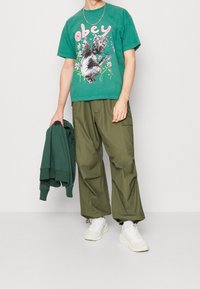 Zelené grafické tričko s nápisem "obey", se vzorem cheruba. Kombinováno s volnými olivovými cargo kalhotami a bílými teniskami.
