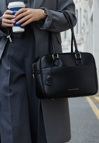 Persona con un traje gris sosteniendo una taza de café azul y un bolso negro de Victoria Hyde London mientras está de pie en una calle.