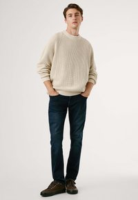 Young man standing with hands in pockets wearing a beige knit sweater, dark blue jeans, and brown casual shoes on white background.