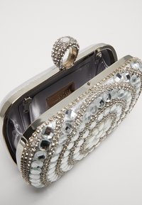 Pochette argentée métallique avec des ornements complexes en cristal, design circulaire et fermoir à anneau orné de pierres précieuses ; intérieur doublé de tissu gris.