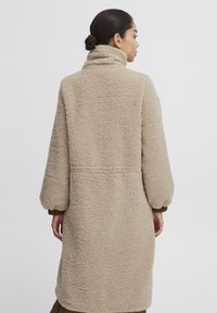 Beige lång kappa med en plysch, texturerad yta, hög krage och överdimensionerade ärmar. Enkel design, sömlös konstruktion och inga synliga knäppningar.